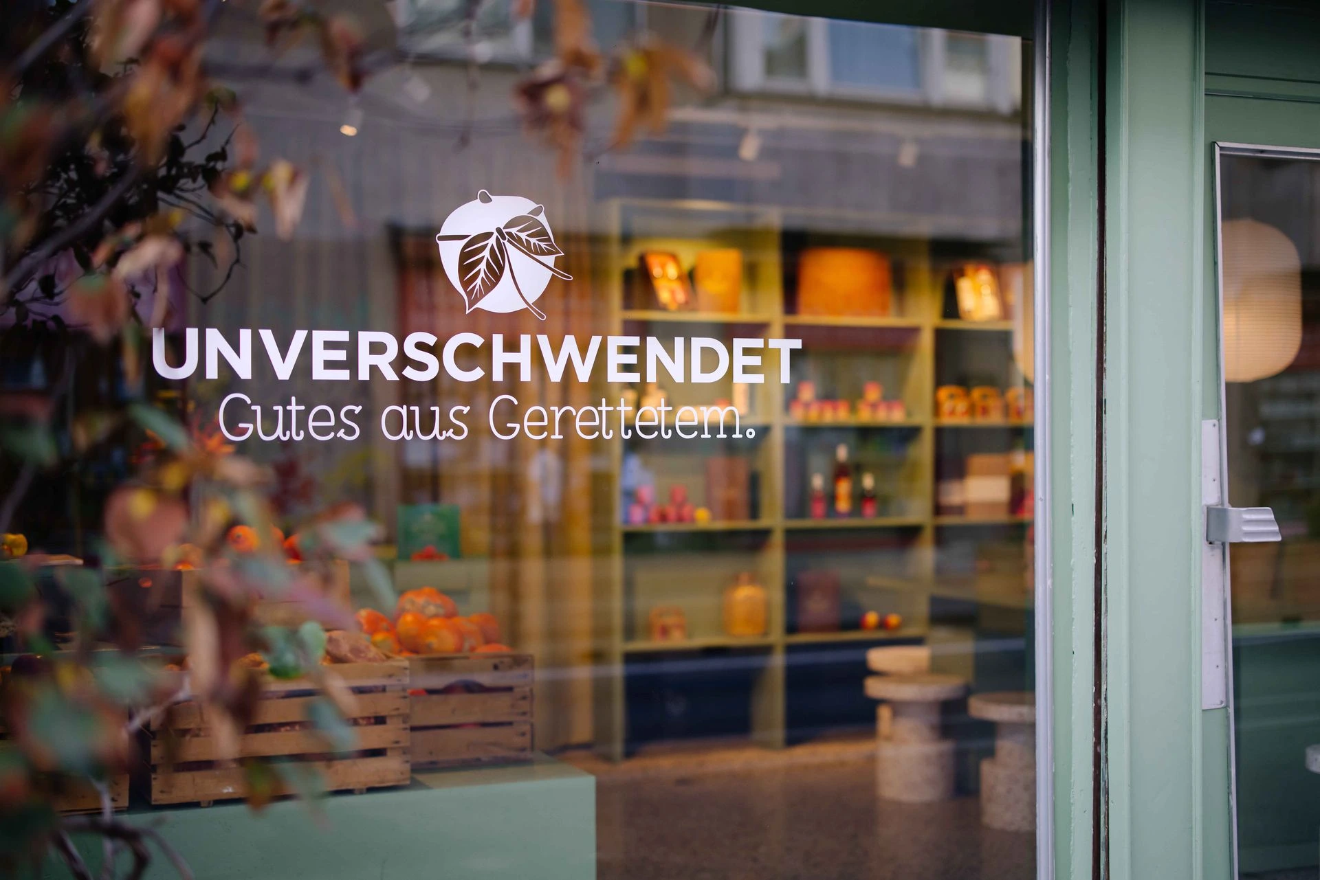 A view through the storefront window of the Unverschwendet shop, showing shelves filled with jars and bottles, and wooden crates containing fresh produce.