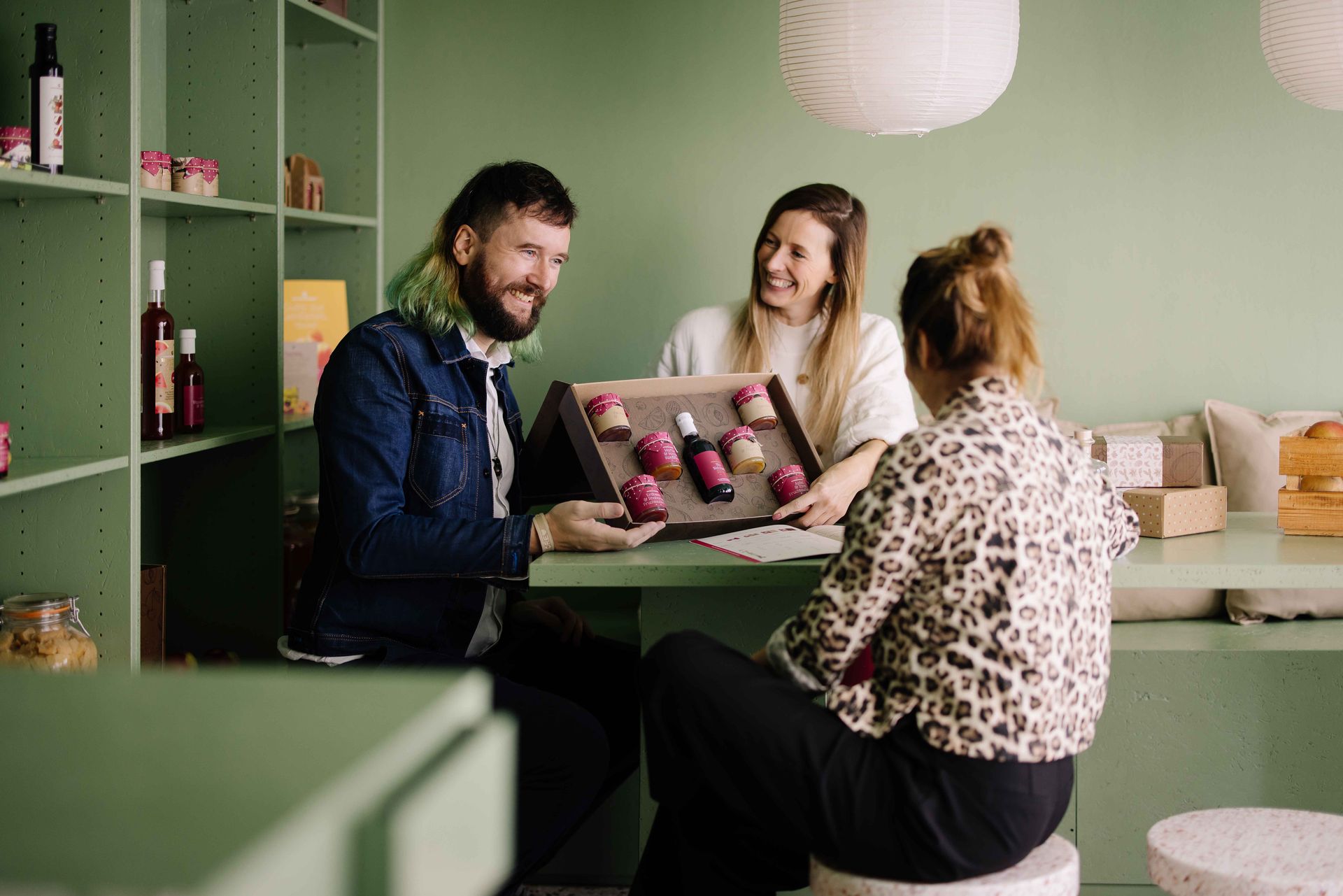 hree people sitting at a green table in a shop, laughing while presenting an open gift box containing various gourmet jars and a bottle.