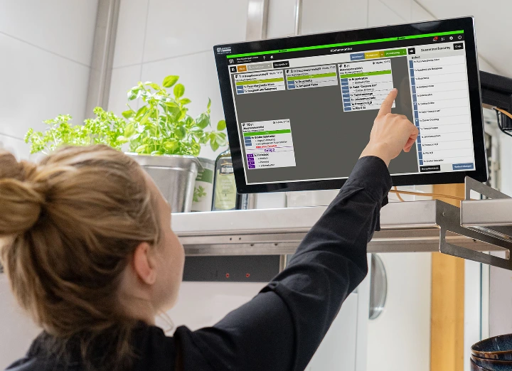 A chef interacting with a Gastronovi digital kitchen monitor system in a professional kitchen, showing real-time order management and workflow automation.